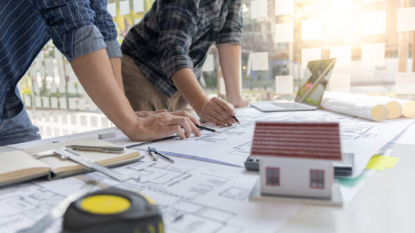 Photo of an architect brainstorming with another person over blueprints on a big table, with a small model house in the foreground.